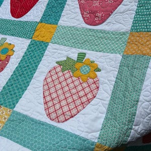 May include: A patchwork quilt featuring white squares with appliqued strawberry motifs in red and pink plaid or floral patterns. Each strawberry is topped with a green leaf and a yellow flower. The squares are separated by teal and yellow fabric strips.