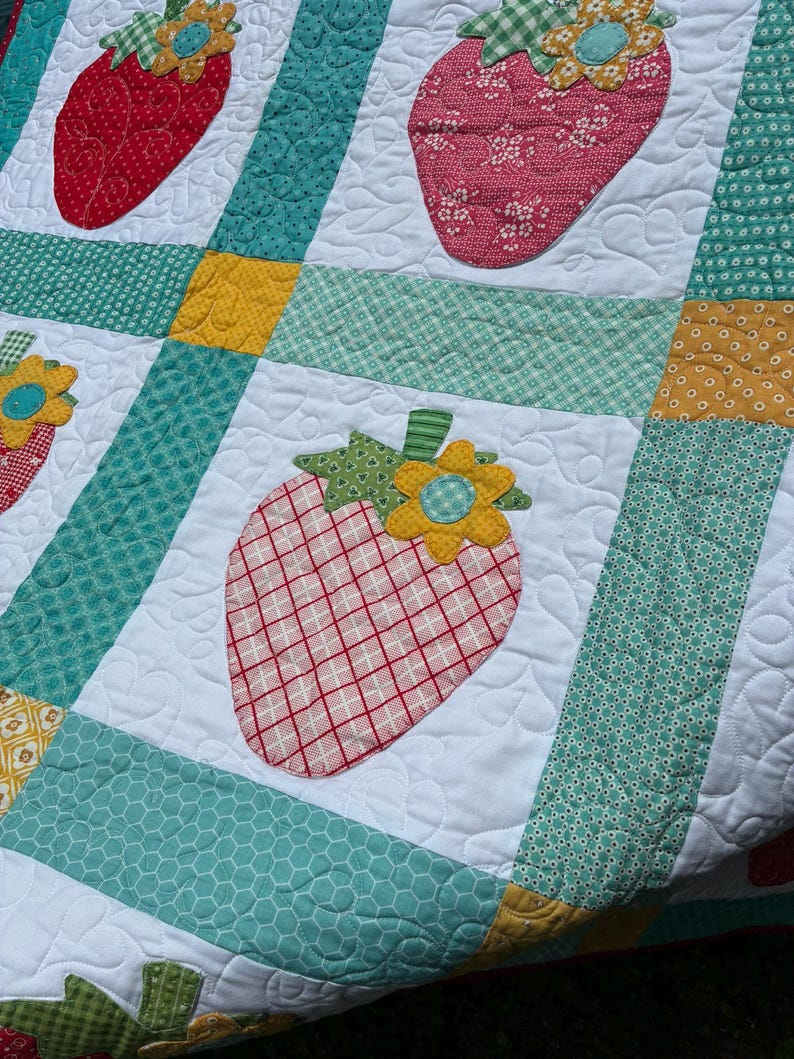 May include: A patchwork quilt featuring white squares with appliqued strawberry motifs in red and pink plaid or floral patterns. Each strawberry is topped with a green leaf and a yellow flower. The squares are separated by teal and yellow fabric strips.