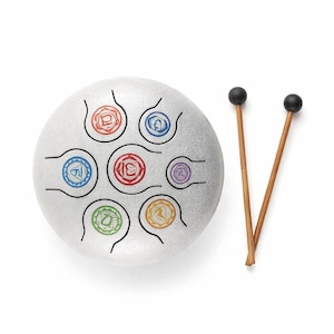 May include: A silver tongue drum with seven colorful chakra symbols. Two wooden drumsticks with black tips are positioned to the right of the drum. The drum is a musical instrument used for meditation and sound healing.