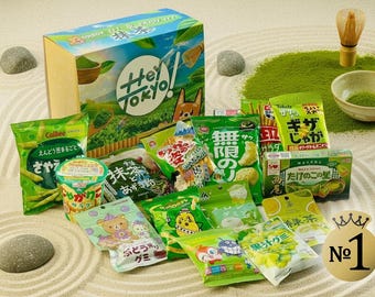 May include: A collection of Japanese snacks, including matcha-flavored items, arranged on a sand-like surface. The packaging features text in Japanese and English, with a box labeled "Hey Tokyo!" and a "No. 1" graphic.