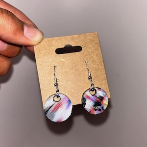 May include: A pair of round earrings with a colorful abstract design. The earrings are attached to silver-colored hooks and are displayed on a brown cardboard backing.