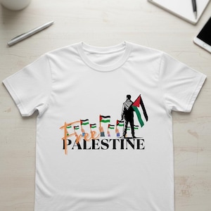 Puede incluir: Camiseta blanca con la frase "Free Palestine" en una fuente estilizada. El diseño incluye la silueta de una persona sosteniendo una bandera palestina, con banderas y manos adicionales. La camiseta está hecha de un material suave y cómodo.