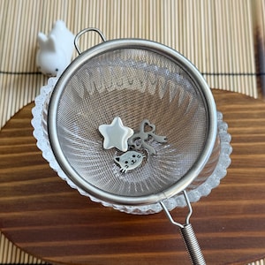 May include: A stainless steel mesh tea strainer with a silver-toned star charm, a silver-toned bow charm, and a silver-toned cat face charm resting inside the bowl. The strainer features a circular rim and a wire handle.