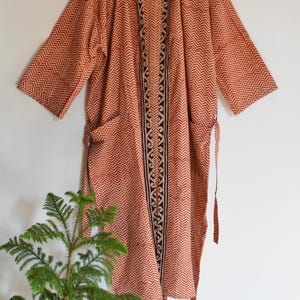 Op de afbeelding: Lange, openvallende kimono in een warme roodbruine tint met een herhalend visgraatpatroon. De kimono heeft een zwart-crèmekleurige decoratieve rand langs de voorkant en mouwen. Het heeft zakken en een striksluiting. De kimono hangt aan een houten hanger.