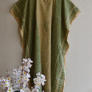 May include: A flowing, beige and green kaftan dress with a V-neck and a geometric pattern. The dress is hanging on a wooden hanger. The dress has a green zigzag pattern and a decorative border. White flowers are in the foreground.