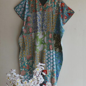 May include: A patchwork caftan in shades of teal, blue, green, and red with floral and geometric patterns. The caftan has a V-neck and short sleeves. White flowers are in the foreground.