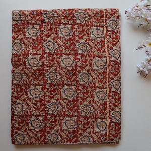 Op de afbeelding: Een gevouwen, rechthoekig textiel met een herhalend bloemenpatroon. Het ontwerp toont grote, gestileerde bloemen in crème en marineblauw tegen een dieprode achtergrond. De stof lijkt gewatteerd met zichtbare stiksels.