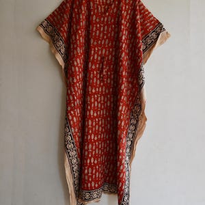 May include: A red caftan with a beige and black patterned border and a repeating white design. The caftan has a V-neck and wide sleeves. It is hanging on a wooden hanger.