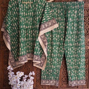 May include: Green two-piece pajama set with a fish pattern. The top has a V-neck and short sleeves. The pants have an elastic waistband. Both pieces have a beige and black patterned border. The set is hanging on a wooden hanger.