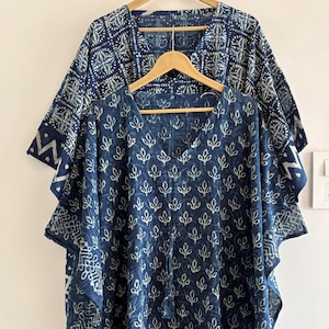 May include: Two indigo blue caftans with white leaf patterns and geometric designs. One caftan is in the foreground with a V-neck and a tassel tie. The other caftan is behind it, with a similar design and a round neckline. Both are hanging on wooden hangers.