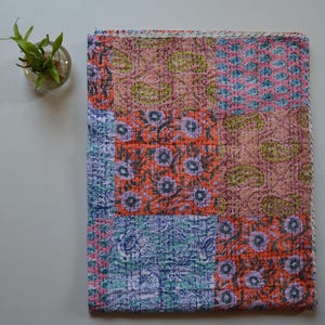 May include: A folded patchwork quilt with a variety of floral and paisley patterns in shades of pink, orange, blue, and green. The quilt is made of stitched fabric and has a white border. A small glass vase with green plants is in the upper left corner.