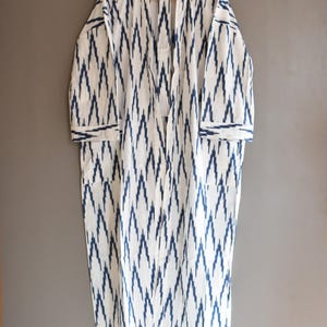 May include: White and blue patterned robe with a V-neck design. The robe features a repeating zigzag pattern in blue against a white background. It has long sleeves and pockets, hanging on a wooden hanger.