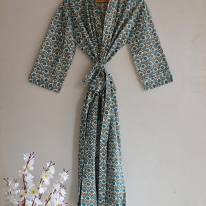 May include: A teal and white patterned robe with a floral design. The robe is tied at the waist and hangs on a wooden hanger. The robe has long sleeves and a white trim. A bouquet of white flowers is in the lower left corner.