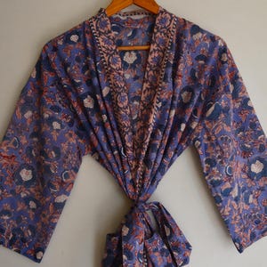 Peut inclure: Robe bleue à motifs floraux avec manches longues. La robe est dotée d'une fermeture à nouer et est suspendue à un cintre en bois. Le motif floral comprend des fleurs et des feuilles blanches, orange et bleues sur fond bleu.