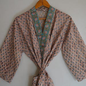 Peut inclure: Une robe rose clair avec un motif floral de petites fleurs bleues et marron. La robe a une bordure turquoise et dorée autour du col et des bords avant. La robe est suspendue à un cintre en bois.