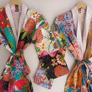 Op de afbeelding: Twee kleurrijke patchwork badjassen hangen aan houten kleerhangers. De badjassen hebben verschillende bloemen- en geometrische patronen in rood, groen, blauw en geel. Elke badjas heeft een witte kraag en een bijpassende strikriem.