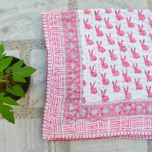 May include: A white and pink patterned quilt featuring a repeating design of pink rabbits. The quilt has a decorative border with floral and geometric patterns. The quilt is folded and placed on a marble surface.