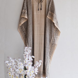 May include: A beige and black patterned kaftan dress. The dress features a geometric design along the edges and a wavy pattern down the center. It has a round neckline with a black tie closure. The dress is hanging on a wooden hanger.