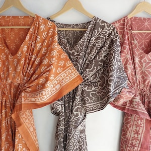 Könnte beinhalten: Drei gemusterte Kaftane in Orange, Braun und Rosa hängen an Holzbügeln. Jeder Kaftan hat einen V-Ausschnitt und eine geraffte Taille. Die Stoffe zeigen sich wiederholende Blumen- und geometrische Muster.