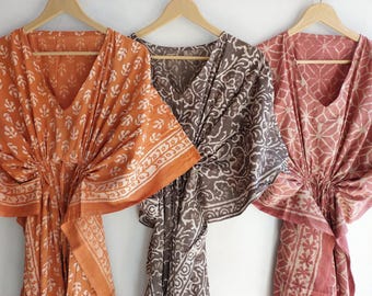Puede incluir: Tres caftanes estampados en tonos naranja, marrón y rosa cuelgan de perchas de madera. Cada caftán presenta un cuello en V y una cintura ceñida. Las telas muestran diseños florales y geométricos repetidos.