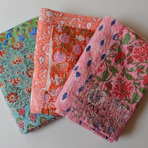 May include: Three folded, quilted fabric items with floral patterns. The fabrics feature a variety of colors, including teal, orange, and pink, with designs of flowers and leaves. The stitching is visible, adding texture to the textiles.