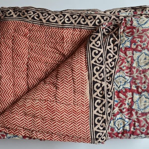 Op de afbeelding: Gevouwen rood-crème quilt met een geometrisch patroon en bloemmotief. De quilt heeft aan één kant een visgraatpatroon en aan de andere kant een bloemenprint. De randen zijn afgewerkt met een zwart-crèmekleurige rand.