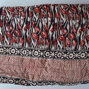 Op de afbeelding: Gevouwen textiel met een bloemenpatroon in rood, zwart en wit. Het bovenste gedeelte heeft een herhalend ontwerp van rode tulpachtige vormen op een zwarte achtergrond met witte stelen. De onderkant heeft een geometrisch patroon in rood en wit.