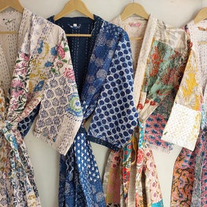 May include: A collection of colorful, patterned robes hanging on wooden hangers. The robes feature floral, geometric, and polka dot designs in various colors, including blue, pink, and yellow. The robes are made of a soft, lightweight fabric.