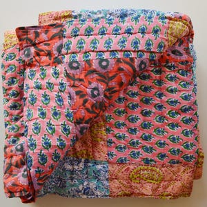 May include: A folded patchwork quilt featuring a variety of floral and paisley patterns in shades of pink, orange, blue, and green. The quilt is made of quilted fabric and has a soft, textured appearance. The design is a mix of different patterns.