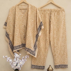 May include: A beige floral print pajama set, including a V-neck top and matching pants, hangs on a wooden hanger. The set features a dark brown border. A vase of white flowers and a decorative lantern are in the foreground.