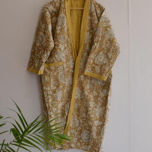 May include: A long, open-front robe with a floral pattern in shades of tan and cream. The robe has a yellow lining and trim, and is hanging on a wooden hanger. The robe is made of a quilted fabric.