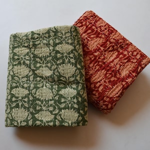 May include: Two folded, quilted textiles. One is green with a floral pattern in cream. The other is red with a floral pattern in tan. Both textiles have visible stitching and a soft, textured appearance.