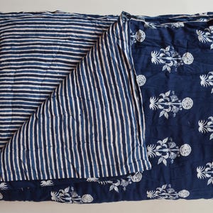 May include: A folded, reversible quilt with a navy blue and white color scheme. One side features horizontal stripes, while the other showcases a floral pattern. The quilt is made of a soft, quilted fabric.