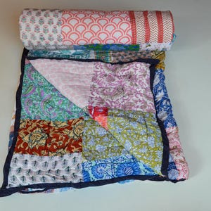 May include: A patchwork quilt featuring a mix of floral and geometric patterns in pink, blue, green, and red. The quilt is folded and rolled, displaying its design and texture. It has a dark blue border. The quilt is made of cotton.
