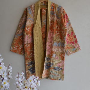 Op de afbeelding: Een patchwork kimono-jas met een bloemenpatroon in tinten oranje, roze en blauw op een beige achtergrond. De jas heeft een los, open ontwerp aan de voorkant en korte mouwen. De binnenvoering is effen beige.