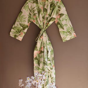 May include: A white robe with a tropical leaf and pink flower pattern. The robe has long sleeves and a tie closure. The robe is hanging on a wooden hanger.