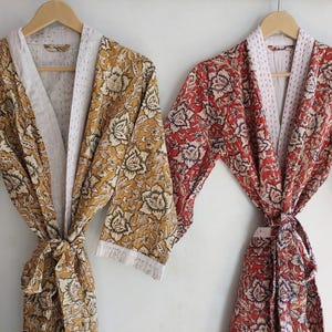 May include: Two floral print robes hanging on wooden hangers. One robe is mustard yellow with white and black floral patterns. The other robe is red with white and blue floral patterns. Both robes have white trim and matching tie belts.