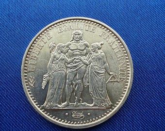 May include: A silver coin featuring a central male figure flanked by two female figures. The coin's edge is detailed, and the words "LIBERTE EGALITE FRATERNITE" are inscribed around the top. The coin has a detailed design and is set against a blue background.
