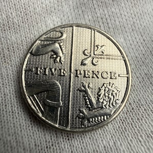 Puede incluir: Una moneda de cinco peniques de plata, con un diseño segmentado con texto e ilustraciones detalladas. La superficie de la moneda tiene un aspecto texturizado, y el texto "FIVE PENCE" se muestra de forma destacada.