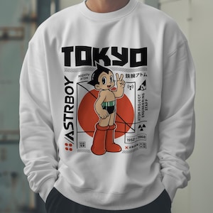 May include: White sweatshirt featuring a graphic of Astro Boy with a peace sign, set against a red circle. The word "TOKYO" is at the top, with Japanese text and "ASTRBOY" on the side. The design includes dates and technical details.