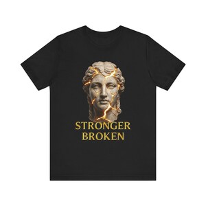 Può includere: T-shirt nero con grafica di un busto classico con crepe dorate e le parole "STRONGER BROKEN" in oro. Il busto ha lineamenti dettagliati e capelli ricci. Il disegno è centrato sulla parte anteriore della maglietta.
