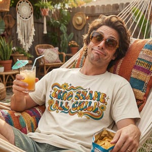 Könnte beinhalten: Beiges T-Shirt mit dem Text "ZERO SHAME ALL CHILL" in Retro-Schrift. Die Person trägt eine Sonnenbrille und entspannt sich in einer Hängematte, hält einen Cocktail mit einem kleinen Schirm und eine Tüte Chips. Szene im Freien.