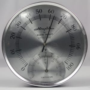 Puede incluir: Un medidor de humedad relativa y temperatura Airguide plateado. El medidor tiene un diseño circular con un dial grande para la humedad y un dial más pequeño para la temperatura en Fahrenheit. La esfera está marcada con números y líneas negras.