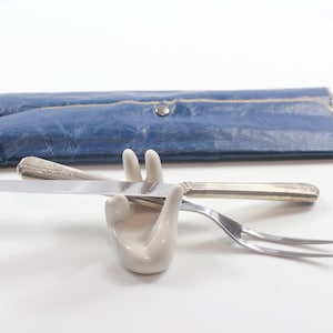 May include: A knife and fork rest on a white surface, held by a ceramic hand-shaped holder. A silver knife and fork are visible. A blue leather case is in the background.