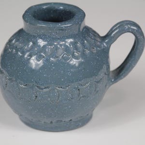 以下が含まれることがあります： 丸みを帯びたボディとハンドルが付いた、斑点のある青いセラミックピッチャー。ピッチャーの中央には装飾模様があり、上部には小さな開口部があります。ピッチャーはおそらく液体を入れるために使用されます。
