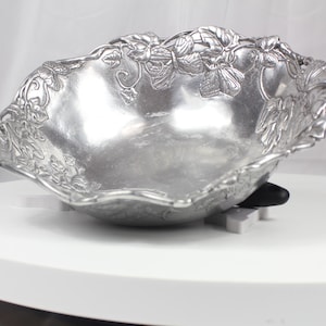 Peut inclure: Un bol décoratif argenté avec un bord festonné et des motifs floraux et de feuilles en relief. Le bol est en métal et repose sur une surface blanche. C'est un objet décoratif.
