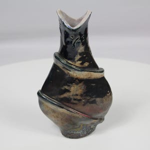 Może przedstawiać: Ceramiczny wazon z błyszczącym czarnym i metalicznie brązowym szkliwem. Naczynie ma asymetryczny, rzeźbiarski kształt ze spiralnie wypukłym pasem owijającym korpus oraz otworem w kształcie litery V u góry, prezentując ciemne, odblaskowe wykończenie.
