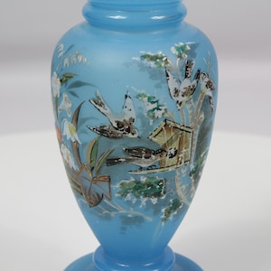 Puede incluir: Un jarrón de vidrio satinado azul claro con una escena pintada a mano de pájaros, una pajarera y flores blancas. El recipiente tiene un cuerpo redondeado, un cuello estrecho y una base ensanchada, con detalles pintados en tonos marrón, blanco y verde.