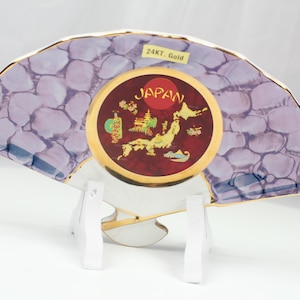 May include: A decorative fan-shaped plate with a purple stone pattern and gold trim. The center features a red circle with "JAPAN" and a gold illustration of a map and figures. The plate is supported by a white stand, and the text "24KT. Gold" is visible.