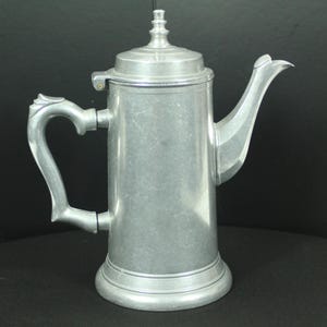 May include: A tall, cylindrical metallic coffee pot with a matte silver finish. It features a hinged lid with a small finial, a curved handle on the left side, and a long, slender spout on the right. The base is slightly flared and circular.